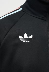 Veste de sport noire à fermeture éclair avec le logo blanc Adidas Trefoil et des rayures pastel rose et bleue sur la couture de l'épaule.