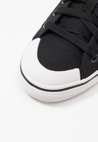 Černé plátěné tenisky s texturovaným černým svrškem, bílým gumovým špičkovým krytem a černými tkaničkami. Mají bílé mezipodešve s černým pruhem.