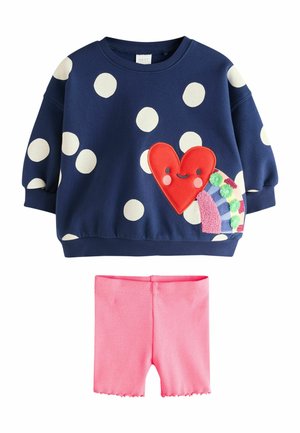 Marineblauer Sweatshirt mit weißen Punkten und einem lächelnden roten Herz-Patch, kombiniert mit rosa gerippten Shorts mit gewellten Kanten.