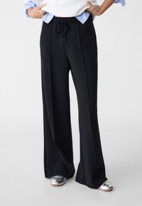 Schwarze weit geschnittene Hose mit elastischem Bund und Kordelzug. Glattes Material, maßgeschneiderter Schnitt und Falten entlang der Vorderseite.