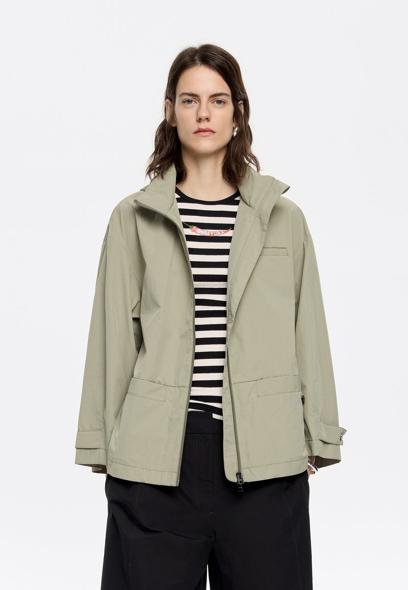 Giacca impermeabile verde chiaro con colletto alto, due tasche frontali e chiusura con zip. Indossata sopra una maglietta a righe bianco e nere.