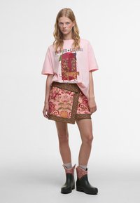 Ροζ γραφικό T-shirt με έντονο σχέδιο, συνδυασμένο με φούστα με λουλουδάτο σχέδιο σε αποχρώσεις ροζ και πράσινου, και μαύρες αδιάβροχες μπότες μέχρι τον αστράγαλο.