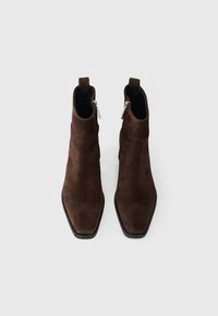 Pár tmavě hnědých semišových kotníkových bot se čtveratými špičkami a bočními zipy, pohled shora na bílém pozadí.