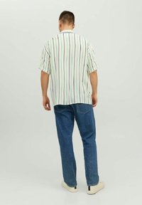 Mężczyzna stojący tyłem, ubrany w białą koszulę z krótkim rękawem w czarne i zielone pionowe paski, niebieskie dżinsy i białe tenisówki.