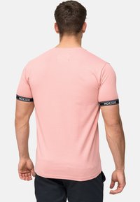 Tricou scurt cu mânecă scurtă de culoare roz deschis, cu guler rotund. Prezintă accente de bandă negre "INDICODE" pe mâneci. Textură de stofă netedă.