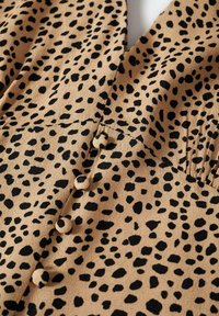 Tela color beige con estampados de animales en negro, con un diseño de escote en V y botones en la parte frontal. Textura suave con un ligero detalle de frunce.