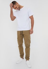 T-shirt blanc à manches courtes, pantalon cargo beige avec poches, et baskets blanches. Le pantalon a des poignets élastiques et une taille avec cordon de serrage.