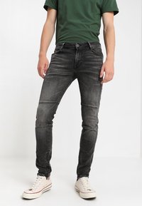 Jeans negros slim fit con textura desgastada, costuras detalladas y un diseño clásico de cinco bolsillos, combinados con zapatillas blancas.