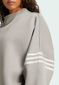 Felpa grigia chiara con collo alto, polsini larghi e strisce bianche sulle maniche. Realizzata in tessuto morbido e testurizzato.