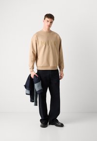 Sweat-shirt beige clair avec logo "HUGO" en relief, associé à un jean noir. Le mannequin tient une veste en denim et porte des chaussures noires. Texture lisse.