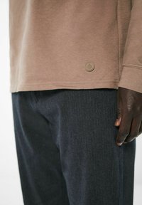 Braunes Langarmhemd mit strukturiertem Rippstoff und einem kleinen runden Logo am Saum, kombiniert mit dunkelblauen Denim-Hosen, die einen dezenten Streifen aufweisen.