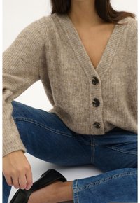 Cardigan beige lavorato a maglia con scollatura a V profonda e tre bottoni scuri. Abbinato a jeans in denim blu e scarpe nere flat. Texture liscia.