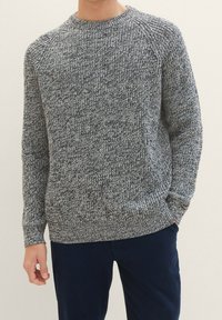 Man in een grijs en zwart gemêleerd gebreid trui met ronde hals en donkerblauwe broek, staande met handen gedeeltelijk in de zakken.