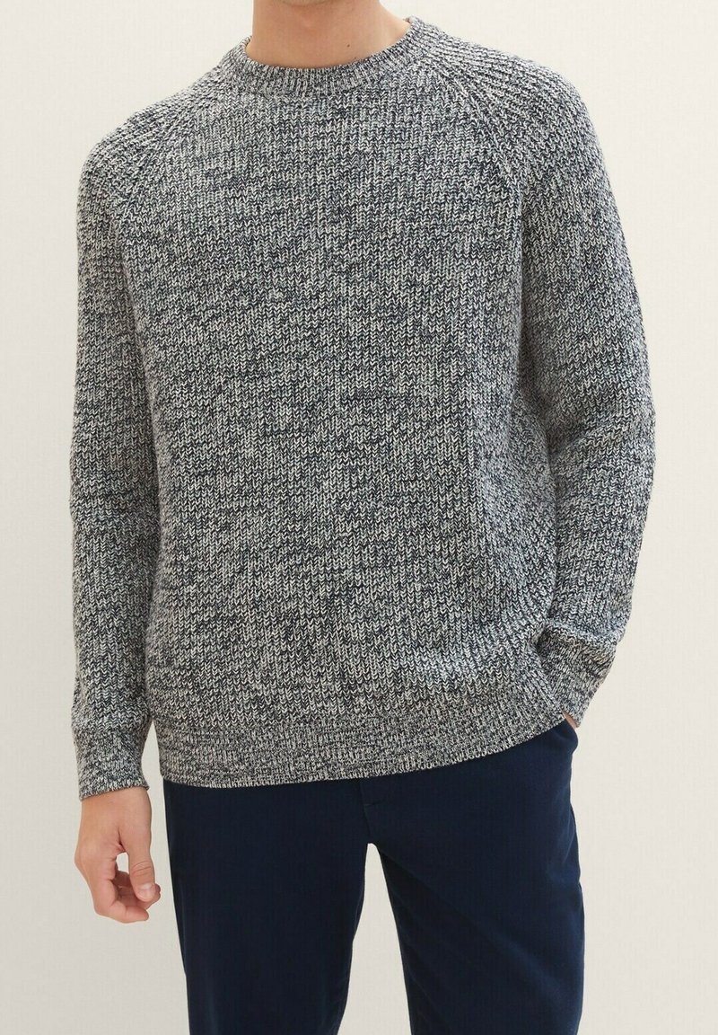 Man in een grijs en zwart gemêleerd gebreid trui met ronde hals en donkerblauwe broek, staande met handen gedeeltelijk in de zakken.