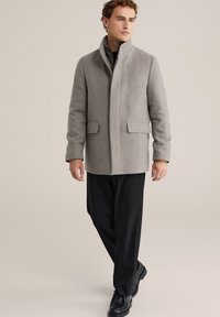 Sivý kabát z vlnenej zmesi s vysokým golierom, dvoma prednými vreckami a minimalistickým dizajnom, skombinovaný s čiernymi nohavicami a čiernymi topánkami.