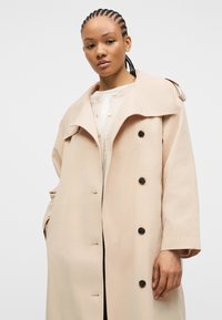 Γυναίκα με πλεξούδες, φορώντας μπεζ διπλόμπουφο καμπαρντίνα και ανοιχτόχρωμη ζακέτα, στέκεται μπροστά σε απλό λευκό φόντο.