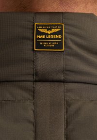 PME Legend oznaka na teksturirani, temno sivi tkanini. Oznaka ima rumene akcentne elemente in besedilo "AMERICAN CLASSIC" ter "TESTED AT HIGH ALTITUDE."
