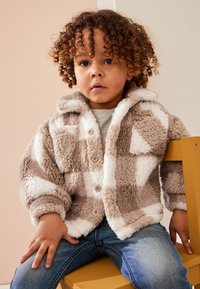Fuzzy, tan og hvid patchwork jakke med hætte; har knaplukninger og blød tekstur. Båret med en grå skjorte og denim jeans.