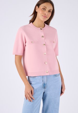 Donna che indossa un cardigan rosa chiaro a maniche corte con bottoni dorati abbinato a jeans azzurri, in piedi di fronte a uno sfondo semplice.