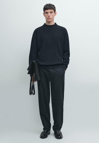 Ciemny sweter dzianinowy z wysokim kołnierzem, w zestawieniu z dopasowanymi szarymi spodniami. Modelka trzyma czarną skórzaną torbę, stoi na neutralnym tle.