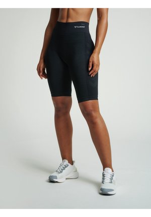 Personne portant un short de sport noir jusqu'au genou et des chaussures de course blanches, debout sur un fond clair uni.