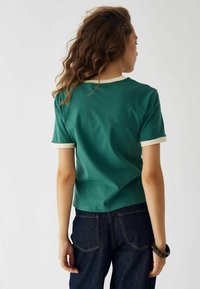 Zelena kratka majica s kremastimi manšetami in ovratnikom, izdelana iz gladkega materiala. Obuta s temnimi jeans hlačami, ki prikazujejo prilega zasnova.