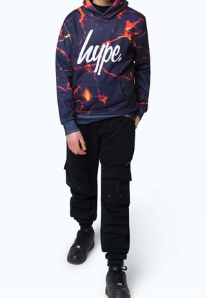 Dziecko w granatowej bluzie z kapturem z pomarańczowym wzorem lawy i białym napisem „hype”, czarne spodnie cargo oraz czarne trampki, stojące na białym tle.