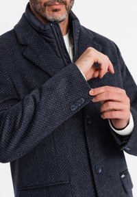Tummansininen teksturoitu takki, jossa on houndstooth-kuvio, korkea kaulus, napitettu hihansuu ja erottuva vuori. Säädettävä istuvuus.
