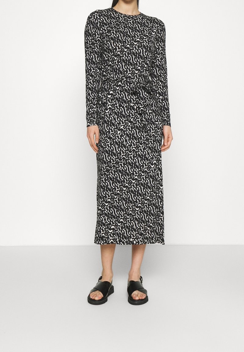 Femme portant une robe midi à manches longues avec un motif abstrait noir et blanc et des sandales noires à bouts ouverts sur un fond uni.