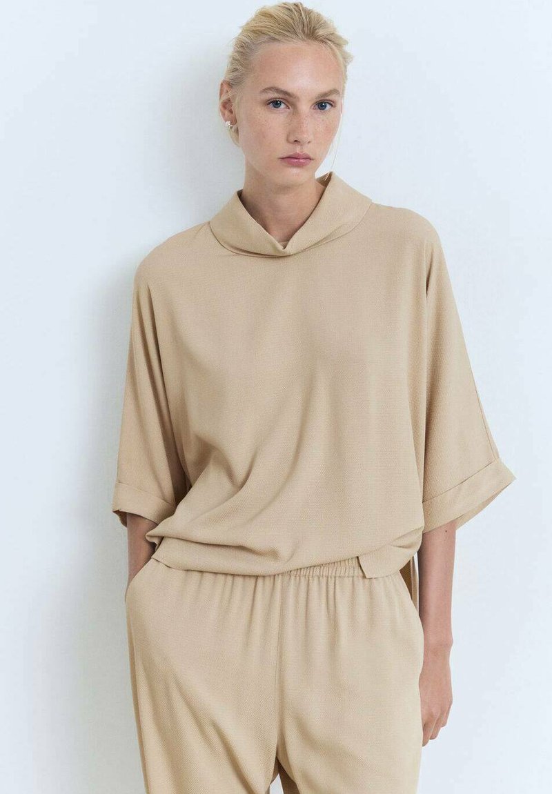 Bež, oversized top s kapuljačom i kratkim rukavima, izrađen od glatkog materijala. Uparen s odgovarajućim hlačama s elastičnim strukom.