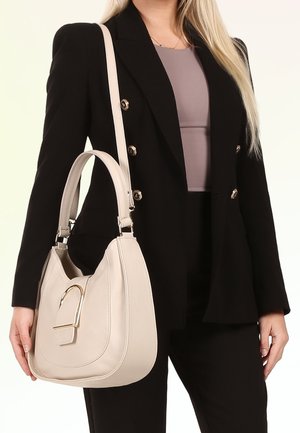 Vrouw draagt een zwarte blazer en broek met een licht mauve top, draagt een crème leren handtas met gouden gesp detail over de schouder.