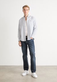 Gestreifte Hemdbluse in Weiß, Blau und Rot, über einem weißen Tanktop getragen. Kombiniert mit dunkelblauen Jeans und weißen Turnschuhen.