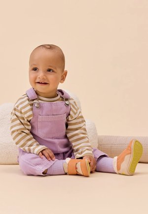Bebé sonriendo sentado en el suelo, vistiendo una camiseta de rayas de manga larga, un overol lavanda, calcetines morados y zapatos suaves de suela beige.
