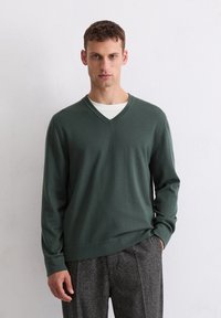 Grüner V-Ausschnitt-Pullover aus weichem Strickmaterial, mit langen Ärmeln und lockerer Passform, kombiniert mit strukturierten grauen Hosen.
