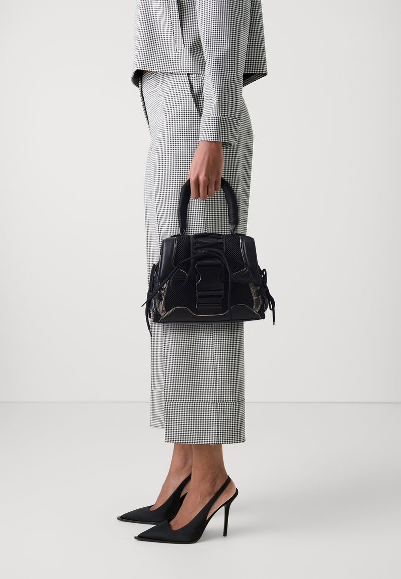 Personne portant un costume à carreaux noir et blanc et des talons noirs, tenant un petit sac à main noir par ses anses.