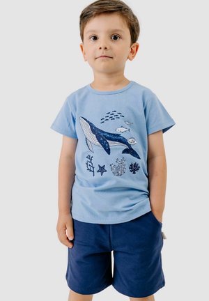 Junge steht mit einer Hand in der Tasche, trägt ein hellblaues T-Shirt mit Wal- und Fischmuster und dunkelblaue Shorts.