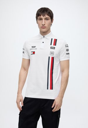Jeune homme portant un polo blanc avec des bandes de course rouges et noires et plusieurs logos de marques automobiles, associé à un pantalon noir.