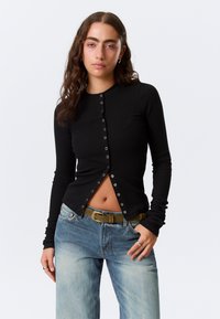 Haut noir à manches longues côtelé avec devant boutonné, col rond et coupe ajustée, associé à un jean en denim bleu clair et une ceinture.