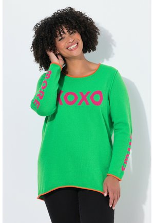 Lächelnde Frau mit lockigem Haar trägt einen leuchtend grünen Pullover mit pinkem "XOXO" auf der Vorderseite und Schriftzügen an den Ärmeln, stehend vor weißem Hintergrund.