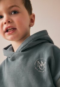 Szara bluza z kapturem o miękkiej teksturze, z białym logo zawierającym tekst "MANHATTAN ISL" i "BIG SMILE CLUB". Zbliżenie na kaptur i dekolt.