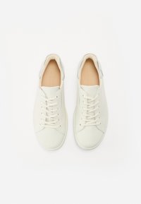 Sneakers in pelle bianca con accenti beige, lacci piatti, design minimalista e dettaglio argento sul tallone, visti dall'alto.