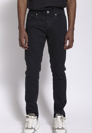 Man met slim-fit zwarte spijkerbroek en witte sneakers, staand tegen een effen lichte achtergrond.