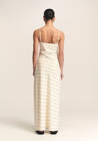 Dolga obleka v kremni barvi s črnimi pikami, tankimi naramnicami, gladkim teksturo in oprijetim zgornjim delom, ki se razteza v krilo do tal.