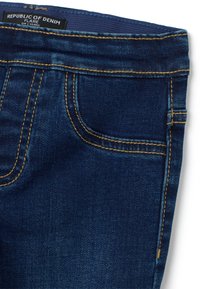 Jeans de mezclilla azul oscuro con un diseño acampanado. Incluye costuras naranjas, una etiqueta en la cinturilla que dice "REPÚBLICA DEL MEZCLO" y bolsillos en la parte frontal.