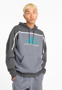 Sudadera gris con mangas en un gris más oscuro. Presenta texto de logo en turquesa "new balance ATHLETIC GEAR" y un bolsillo frontal. Tejido suave.