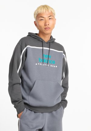 Sudadera gris con mangas en un gris más oscuro. Presenta texto de logo en turquesa "new balance ATHLETIC GEAR" y un bolsillo frontal. Tejido suave.