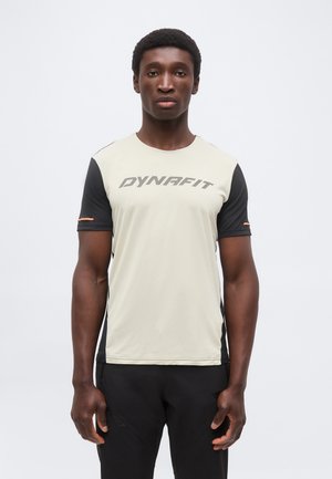 Bărbatul stă cu fața înainte, purtând un tricou sport bej și negru cu mâneci scurte, cu inscripția „DYNAFIT” pe piept și pantaloni negri.