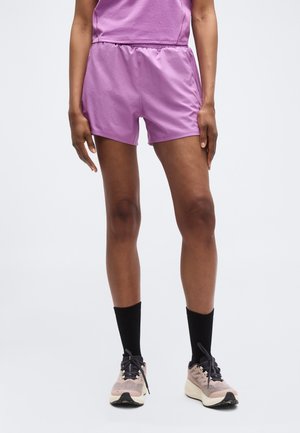 Άτομο που φοράει ανοιχτό μωβ αθλητικές βερμούδες, ταιριαστό τοπ, μαύρες κάλτσες μέχρι τη μέση της γάμπας και μπεζ παπούτσια τρεξίματος σε λευκό φόντο.