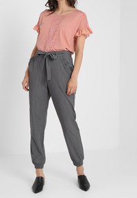 Kvinna som bär en rosa kortärmad topp med spets och volanger, grå högmidjade papperspåsebyxor med en knytbälte, och svarta spetsiga skor.