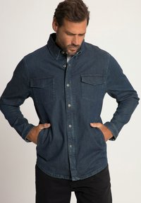 Camicia di denim blu scuro con chiusura a bottoni frontale, due tasche sul petto e colletto a punta, caratterizzata da una texture liscia e maniche lunghe.